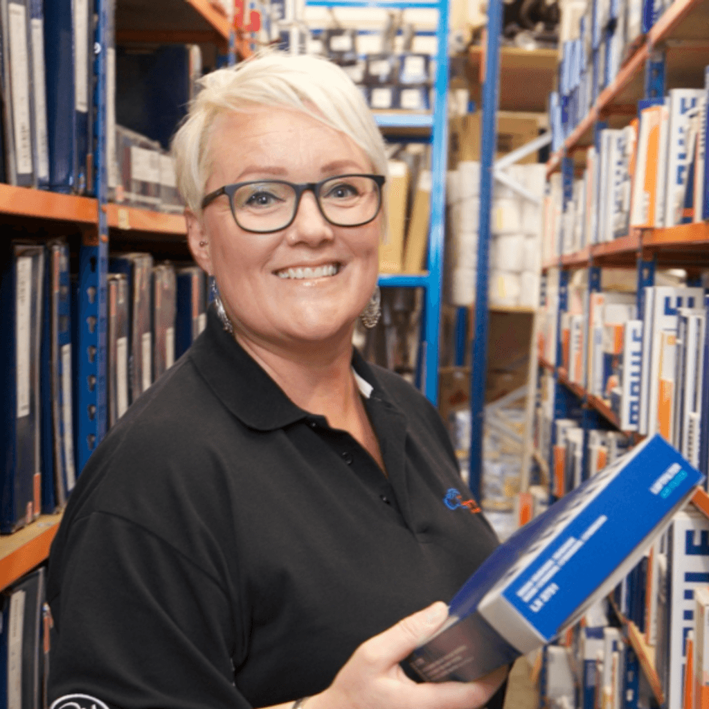Caucasian lady standing in the middle of an aisle holding a book. There are collections of books on both sides.